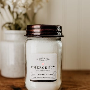 May include: A clear glass jar candle with a dark brown lid. The label reads "EMERGENCY" and "Rosemary & Citrus". The candle is 12 oz. and is from The Driftwood Home.