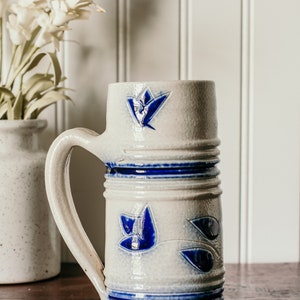 May include: A white ceramic mug with a blue floral design and a blue rim. The mug has a handle and is sitting on a wooden surface.