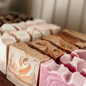 May include: A variety of handmade soap bars in different colors and patterns. The bars are arranged in rows on a wooden surface. The soap bars are made with natural ingredients and are perfect for all skin types.