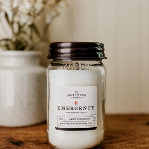 May include: A clear glass jar candle with a dark brown lid. The label reads "The Driftwood Home" and "Emergency" with a plus sign. The candle is 12 oz and scented "Sweet Peppermint".