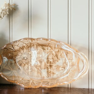 May include: A vintage glass serving dish with a delicate floral pattern. The dish is made of clear glass with a light pink hue and features a scalloped edge. It has two handles on the sides.