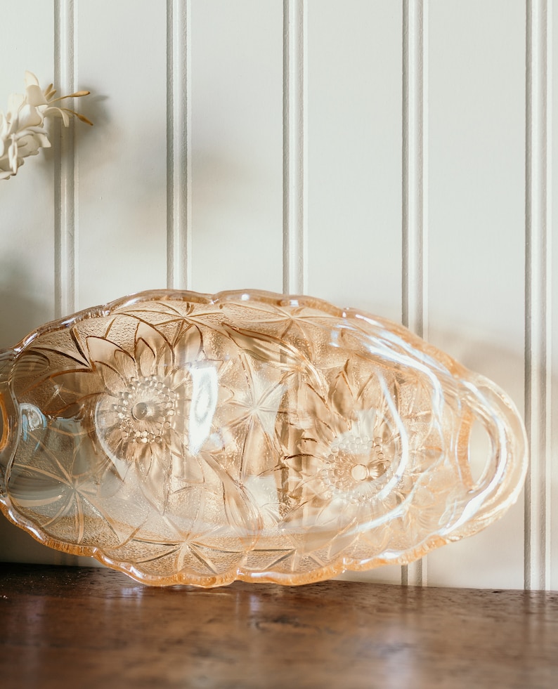 May include: A vintage glass serving dish with a delicate floral pattern. The dish is made of clear glass with a light pink hue and features a scalloped edge. It has two handles on the sides.