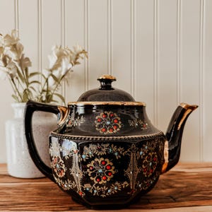 May include: A black ceramic teapot featuring a gold-trimmed lid and spout, decorated with intricate gold filigree patterns, small red circular accents, and tiny light blue dots arranged in a repeating floral motif around the body.