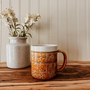 May include: A ceramic mug with a white glazed rim and a brown body featuring embossed geometric, fish, leaf, and mask patterns. Beside the mug sits a speckled white ceramic vase containing white flowers, all set against a vertical paneled wall.