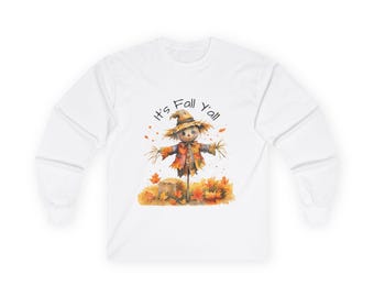 May include: White long-sleeve shirt featuring a watercolor illustration of a scarecrow with a straw hat and autumn leaves. The text "It's Fall Y'all" is above the scarecrow. The design has orange, brown, and yellow colors.