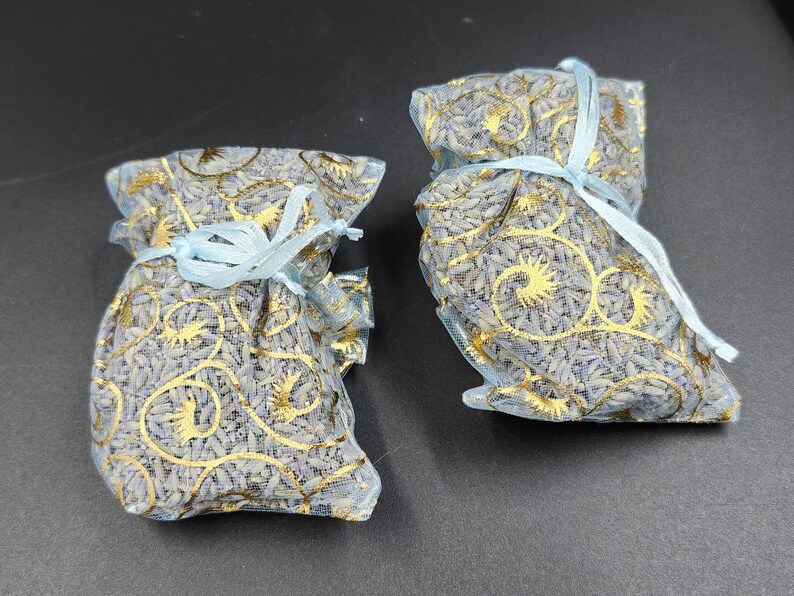 Puede incluir: Dos peque&ntilde;os saquitos de malla llenos de capullos de lavanda. Los saquitos son de color azul claro con dise&ntilde;os dorados y est&aacute;n atados con cintas azul claro. El fondo es una superficie gris oscuro.