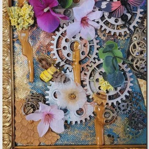 May include: A mixed media wall art piece in a gold ornate frame featuring silver-toned mechanical gears, artificial purple and white flowers, a small yellow and black bee figurine, a butterfly accent, and textured honeycomb patterns on a blue background.
