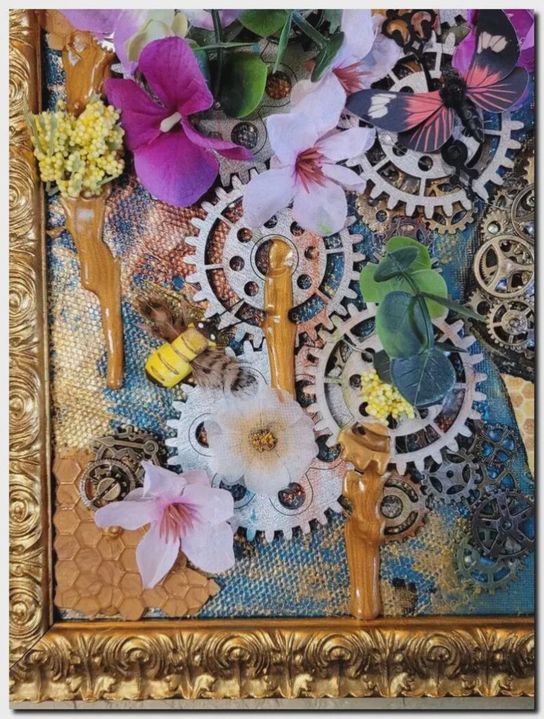 May include: A mixed media wall art piece in a gold ornate frame featuring silver-toned mechanical gears, artificial purple and white flowers, a small yellow and black bee figurine, a butterfly accent, and textured honeycomb patterns on a blue background.