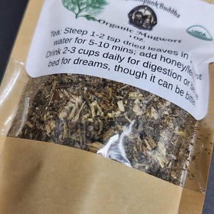 May include: A brown paper bag with a clear window, filled with dried herbs. The label reads "Organic Mugwort" and includes instructions for steeping the tea. The Steampunk Buddha logo is visible.