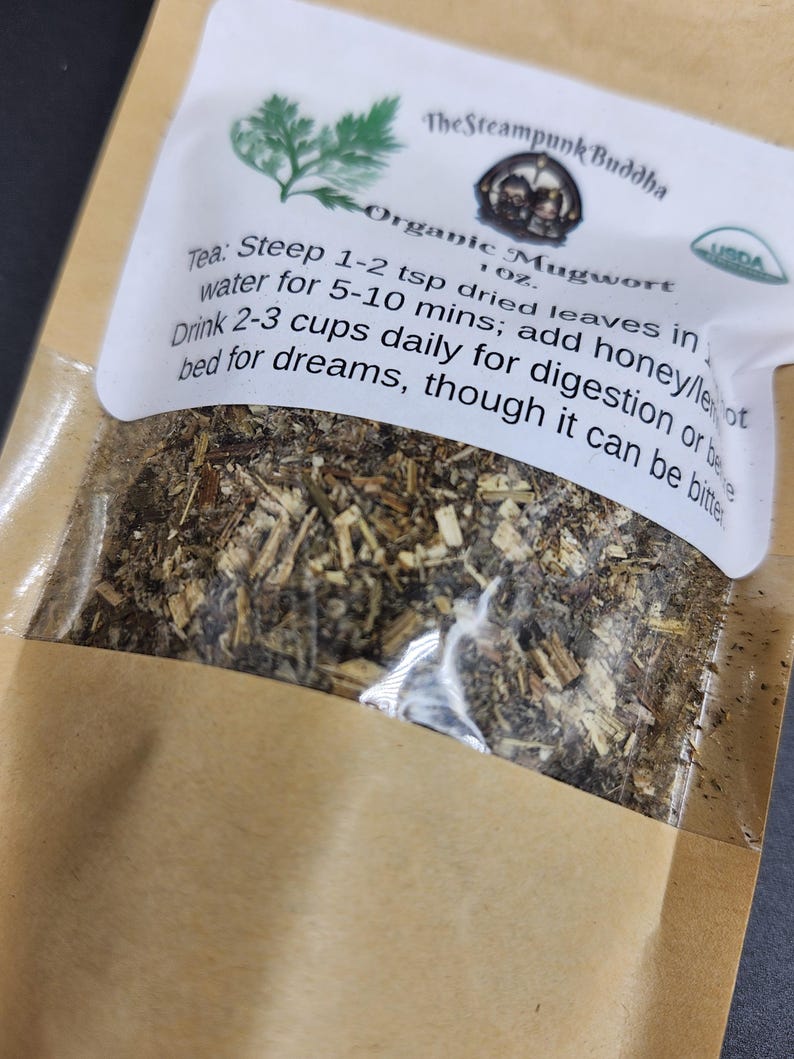 May include: A brown paper bag with a clear window, filled with dried herbs. The label reads "Organic Mugwort" and includes instructions for steeping the tea. The Steampunk Buddha logo is visible.