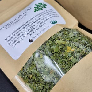 Op de afbeelding: Een transparante zak met gedroogde moringabladthee. Het etiket leest "MORINGA LEAF 1 OZ DRIED HERB" en bevat instructies voor het zetten van thee en het USDA Organic-logo. De zak is gevuld met groene bladeren.
