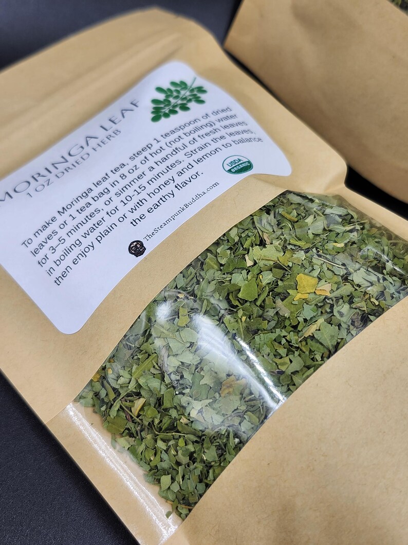 Op de afbeelding: Een transparante zak met gedroogde moringabladthee. Het etiket leest "MORINGA LEAF 1 OZ DRIED HERB" en bevat instructies voor het zetten van thee en het USDA Organic-logo. De zak is gevuld met groene bladeren.