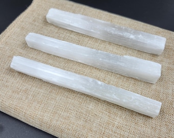 May include: Three white selenite crystal wands, each with a rectangular shape and a slightly textured surface. The wands are arranged horizontally on a tan burlap cloth, creating a natural and earthy aesthetic.