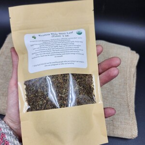 May include: A package of Krishna Holy Basil Leaf (Tulsi) 1 oz. The brown paper bag has a clear window showing dried basil leaves. Text on the label describes the herb's uses for prosperity, love, and mental clarity. The package is held by a person.