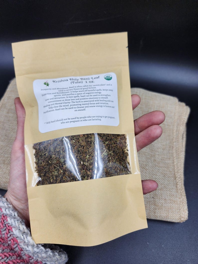 May include: A package of Krishna Holy Basil Leaf (Tulsi) 1 oz. The brown paper bag has a clear window showing dried basil leaves. Text on the label describes the herb's uses for prosperity, love, and mental clarity. The package is held by a person.