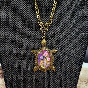 K&ouml;nnte beinhalten: Eine bronzefarbene Schildkr&ouml;ten-Halskette mit einer Kette und einem schmetterlingsf&ouml;rmigen Verbindungsst&uuml;ck. Der Schildkr&ouml;tenpanzer enth&auml;lt eine violette ovale Einlage, die mit kleinen bunten Blumenformen und metallischen Zahnr&auml;dern verziert ist. Der Text TheSteampunkBuddha ist sichtbar.