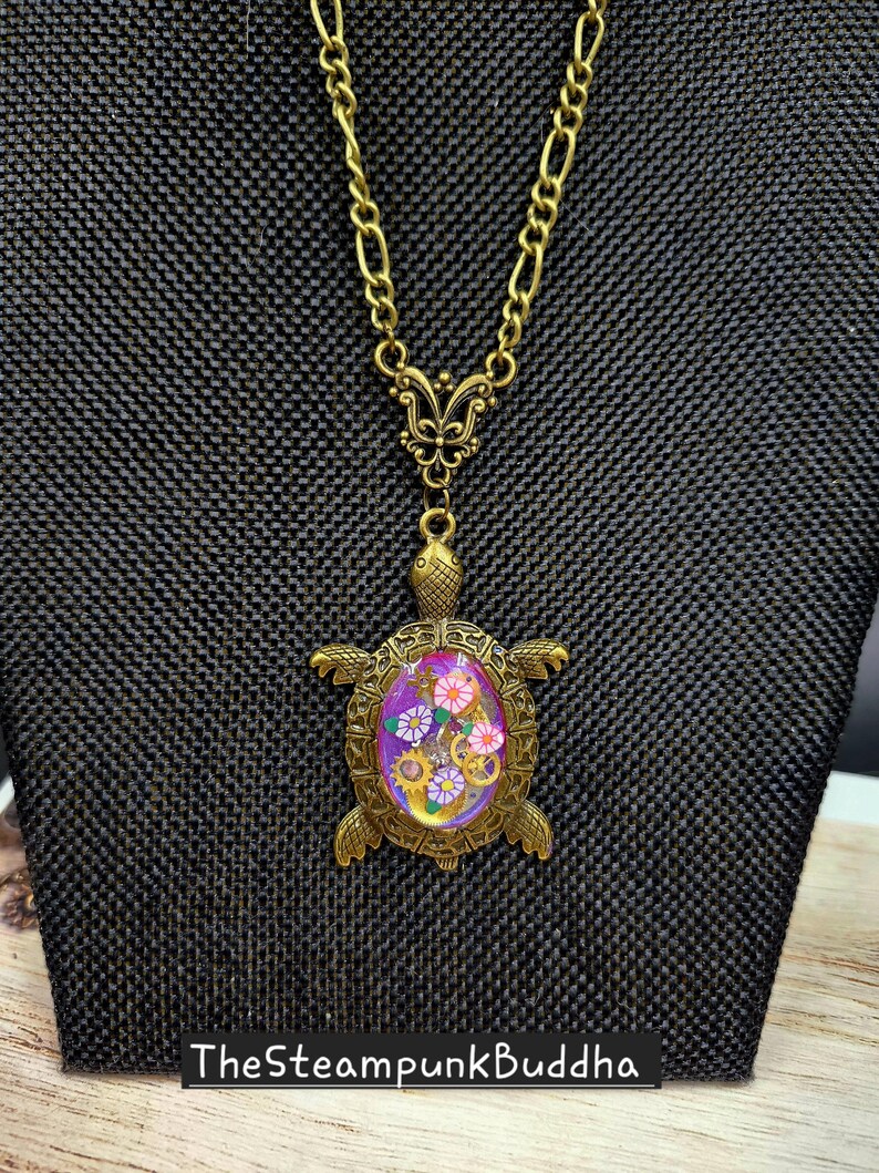 K&ouml;nnte beinhalten: Eine bronzefarbene Schildkr&ouml;ten-Halskette mit einer Kette und einem schmetterlingsf&ouml;rmigen Verbindungsst&uuml;ck. Der Schildkr&ouml;tenpanzer enth&auml;lt eine violette ovale Einlage, die mit kleinen bunten Blumenformen und metallischen Zahnr&auml;dern verziert ist. Der Text TheSteampunkBuddha ist sichtbar.