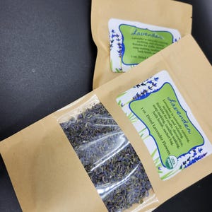 以下が含まれることがあります： 透明な窓が付いた2つの茶色の紙袋。乾燥ラベンダーの花が詰まっています。各バッグには「ラベンダー」と書かれたラベルがあり、石鹸作りやキャンドル作りなどの用途が記載されています。バッグには乾燥ラベンダーの花が28グラム入っています。
