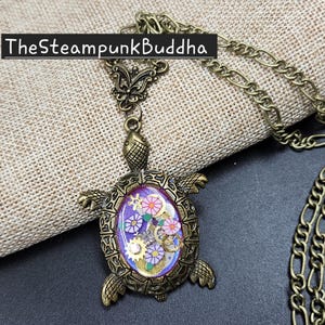 K&ouml;nnte beinhalten: Eine bronzefarbene Schildkr&ouml;ten-Halskette mit einem violetten ovalen Zentrum, das mit kleinen goldfarbenen Zahnr&auml;dern und rosa blumenf&ouml;rmigen Akzenten gef&uuml;llt ist. Die Schildkr&ouml;te hat einen strukturierten Metallpanzer und Kopf, Text TheSteampunkBuddha sichtbar.