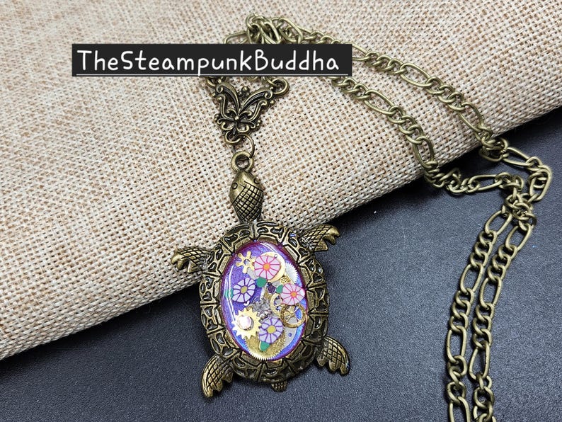K&ouml;nnte beinhalten: Eine bronzefarbene Schildkr&ouml;ten-Halskette mit einem violetten ovalen Zentrum, das mit kleinen goldfarbenen Zahnr&auml;dern und rosa blumenf&ouml;rmigen Akzenten gef&uuml;llt ist. Die Schildkr&ouml;te hat einen strukturierten Metallpanzer und Kopf, Text TheSteampunkBuddha sichtbar.