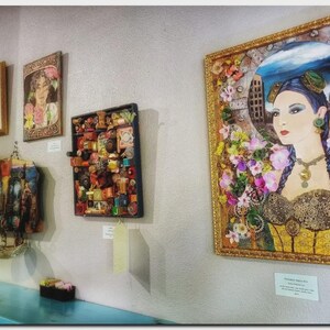 May include: A collection of framed wall art pieces displayed on a wall, including a large portrait of a person with blue hair and floral accents, a square assemblage of small objects, and two smaller framed prints above a hanging textile piece.
