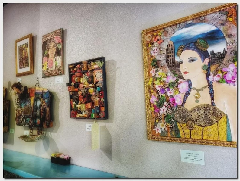 May include: A collection of framed wall art pieces displayed on a wall, including a large portrait of a person with blue hair and floral accents, a square assemblage of small objects, and two smaller framed prints above a hanging textile piece.