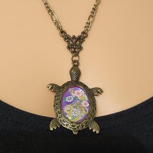 K&ouml;nnte beinhalten: Eine bronzefarbene Schildkr&ouml;ten-Halskette mit einem ovalen, violetten Inlay. Der Panzer der Schildkr&ouml;te ist mit kleinen metallischen Zahnr&auml;dern und kreisf&ouml;rmigen Elementen mit Blumenmuster verziert. Der Anh&auml;nger h&auml;ngt an einer passenden Kette.