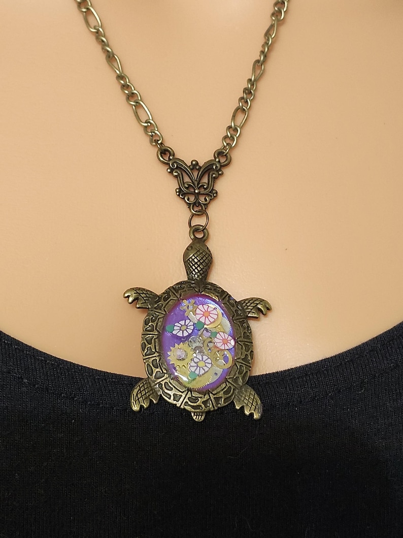K&ouml;nnte beinhalten: Eine bronzefarbene Schildkr&ouml;ten-Halskette mit einem ovalen, violetten Inlay. Der Panzer der Schildkr&ouml;te ist mit kleinen metallischen Zahnr&auml;dern und kreisf&ouml;rmigen Elementen mit Blumenmuster verziert. Der Anh&auml;nger h&auml;ngt an einer passenden Kette.