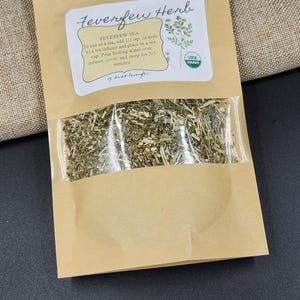 Puede incluir: Una bolsa de papel marr&oacute;n con una ventana transparente que muestra hierba de matricaria seca. La bolsa tiene una etiqueta con el texto "Feverfew Herb" e instrucciones para usarla como t&eacute;. Se ve un sello org&aacute;nico USDA.