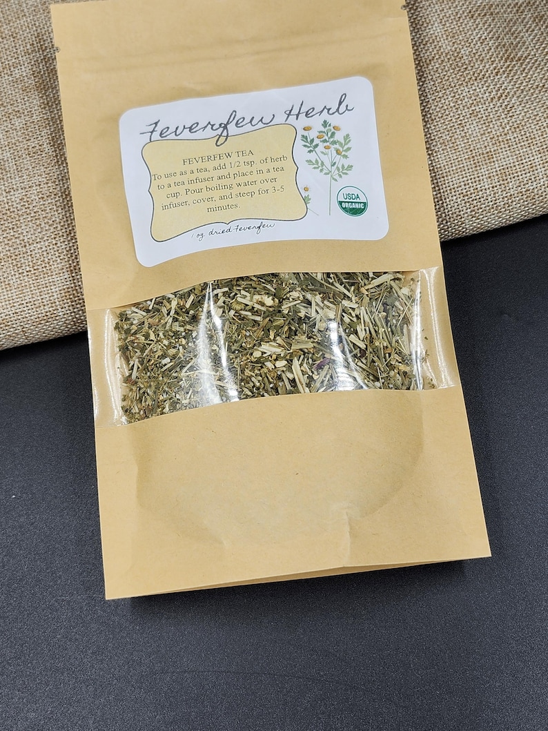Puede incluir: Una bolsa de papel marr&oacute;n con una ventana transparente que muestra hierba de matricaria seca. La bolsa tiene una etiqueta con el texto "Feverfew Herb" e instrucciones para usarla como t&eacute;. Se ve un sello org&aacute;nico USDA.
