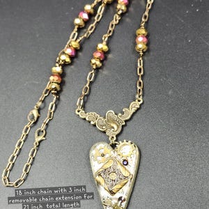 May include: A steampunk-style necklace with an 18-inch chain and a 3-inch extension for a total length of 21 inches. The pendant is heart-shaped, featuring gears, a dragonfly, and a decorative design. The chain has gold and pink beads.