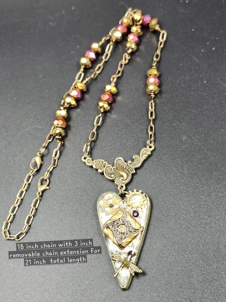 May include: A steampunk-style necklace with an 18-inch chain and a 3-inch extension for a total length of 21 inches. The pendant is heart-shaped, featuring gears, a dragonfly, and a decorative design. The chain has gold and pink beads.