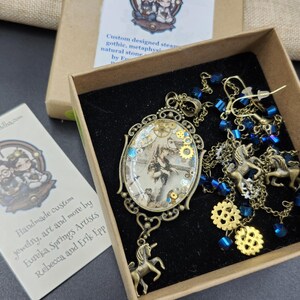 May include: A steampunk-themed jewelry set in a box. The set includes a pendant with gears and a unicorn charm, earrings, and a necklace with blue beads and unicorn charms. The box contains a card with the text "Handmade custom jewelry, art and more by Eureka Springs Artists Rebecca and Erik Epp."