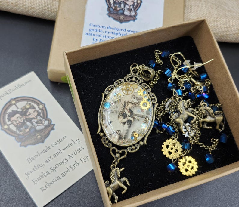 May include: A steampunk-themed jewelry set in a box. The set includes a pendant with gears and a unicorn charm, earrings, and a necklace with blue beads and unicorn charms. The box contains a card with the text "Handmade custom jewelry, art and more by Eureka Springs Artists Rebecca and Erik Epp."