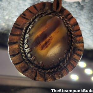 K&ouml;nnte beinhalten: Handgefertigter Anh&auml;nger mit einem ovalen Stein in Braun- und Gelbt&ouml;nen. Der Stein ist von einem detaillierten, strukturierten Metallrand mit Seilmotiv umgeben. Der Anh&auml;nger hat eine Steampunk-&Auml;sthetik, mit dem Text "TheSteampunkBuddha" unten.