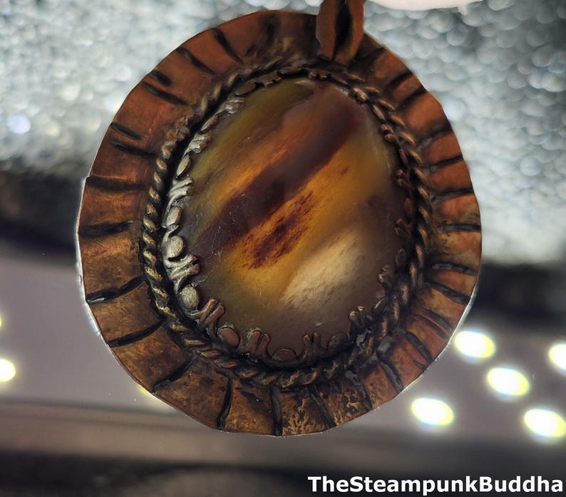 K&ouml;nnte beinhalten: Handgefertigter Anh&auml;nger mit einem ovalen Stein in Braun- und Gelbt&ouml;nen. Der Stein ist von einem detaillierten, strukturierten Metallrand mit Seilmotiv umgeben. Der Anh&auml;nger hat eine Steampunk-&Auml;sthetik, mit dem Text "TheSteampunkBuddha" unten.