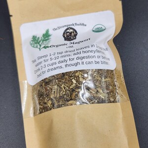 May include: A brown paper bag with a clear window containing dried herbs. The label reads "Organic Mugwort" and includes instructions for use. The bag is sealed and has a USDA organic seal.