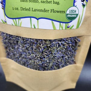 以下が含まれることがあります： 乾燥ラベンダーの花が入った茶色の紙袋のクローズアップ。袋には「1 oz. Dried Lavender Flowers USDA ORGANIC」と書かれた緑色のラベルが付いています。ラベンダーは紫と緑の混合です。