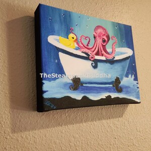 May include: Square canvas art featuring a pink octopus in a blue bathtub with a yellow rubber duck. The artwork has a whimsical style, with water droplets and the text "TheSteampunkBuddha" visible. The canvas is mounted on a wooden frame.