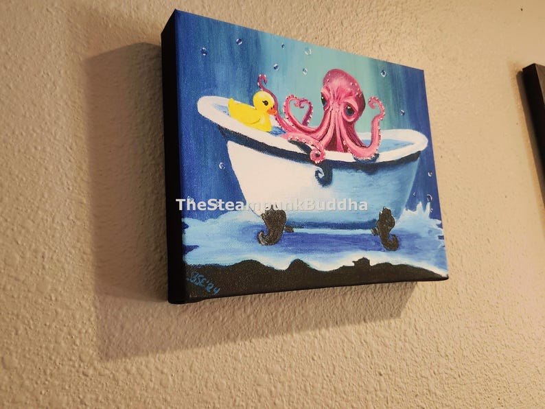 May include: Square canvas art featuring a pink octopus in a blue bathtub with a yellow rubber duck. The artwork has a whimsical style, with water droplets and the text "TheSteampunkBuddha" visible. The canvas is mounted on a wooden frame.