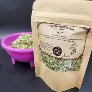 May include: A brown paper pouch labeled The Steampunk Buddha Rue containing dried green herbs, with a small pink bowl filled with the same dried herbs positioned to the left. The label includes brewing directions and a small cartoon illustration.