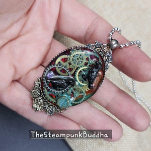 Peut inclure: Pendentif ovale avec un design steampunk. Le pendentif pr&eacute;sente des engrenages, un oiseau noir et un accent turquoise, sertis dans une mati&egrave;re semblable &agrave; de la r&eacute;sine. Le texte "TheSteampunkBuddha" est visible en bas.