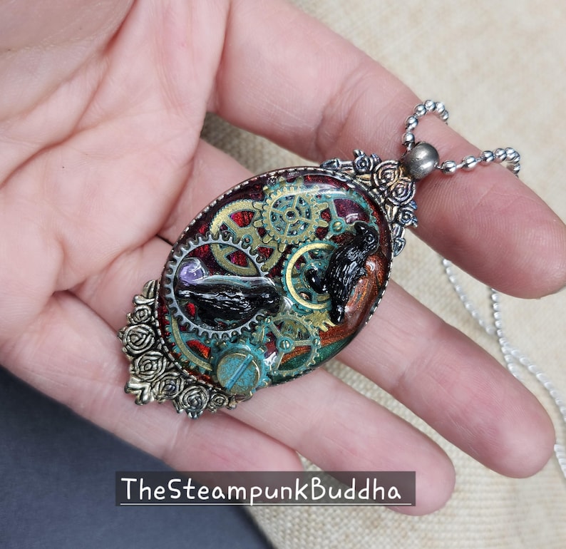 Peut inclure: Pendentif ovale avec un design steampunk. Le pendentif pr&eacute;sente des engrenages, un oiseau noir et un accent turquoise, sertis dans une mati&egrave;re semblable &agrave; de la r&eacute;sine. Le texte "TheSteampunkBuddha" est visible en bas.