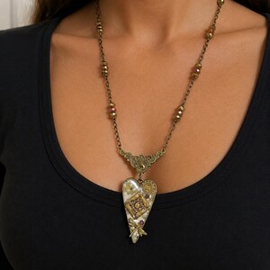 May include: A necklace featuring a heart-shaped pendant with a cream-colored background, gold-toned mechanical gear accents, and a small dragonfly charm. The pendant hangs from a bronze-colored chain with faceted beads integrated into the links.
