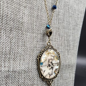 May include: Antique brass-toned necklace featuring a pendant with a vintage illustration. The oval pendant showcases a black and white image with gold and blue accents. The necklace chain includes blue crystal beads and a small unicorn charm.