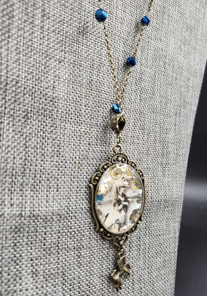 May include: Antique brass-toned necklace featuring a pendant with a vintage illustration. The oval pendant showcases a black and white image with gold and blue accents. The necklace chain includes blue crystal beads and a small unicorn charm.