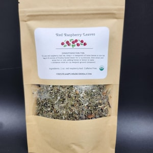 May include: A brown paper bag with a clear window, filled with dried herbs. The label reads "Red Raspberry Leaves" with instructions for tea preparation. The bag contains 1 oz of red raspberry leaf and is caffeine-free. The website address is visible.