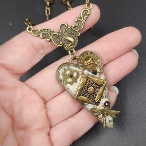 May include: A steampunk-style heart pendant necklace in antique brass. The heart features gears, filigree, and a dragonfly charm. The pendant is approximately 2 inches long and hangs from a delicate chain.