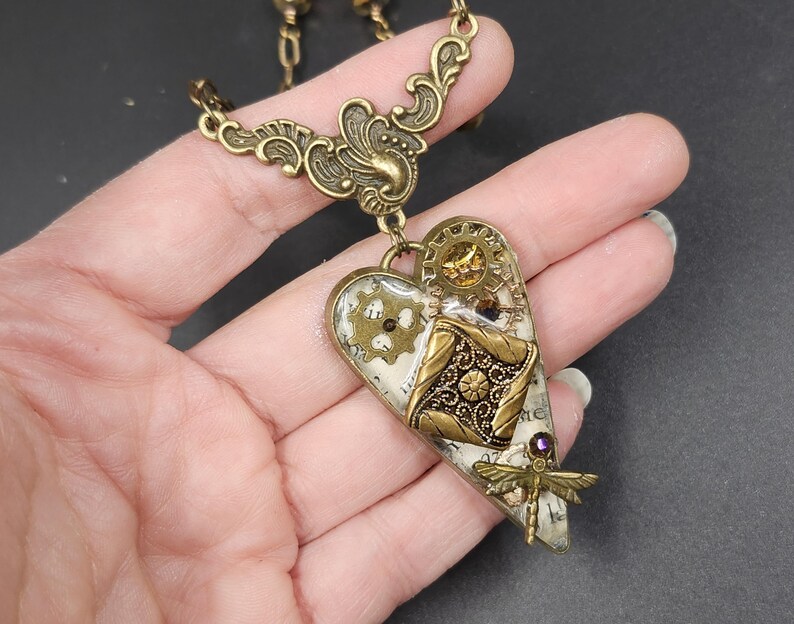 May include: A steampunk-style heart pendant necklace in antique brass. The heart features gears, filigree, and a dragonfly charm. The pendant is approximately 2 inches long and hangs from a delicate chain.