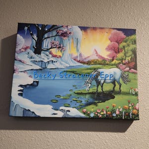 Puede incluir: Una pintura sobre lienzo representa un paisaje sereno con un unicornio blanco pastando cerca de un estanque. La obra de arte presenta una mezcla de paisajes invernales y primaverales, con nieve, hielo y flores floreciendo. El nombre del artista, "Becky Stracener Epp", es visible.